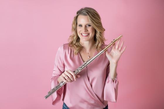 Julianna Nickel, a white woman with short wavy blond hair, stands in front of a pink background, smiling at the camera and holding her flute in her left hand. Nickel is an adjunct professor at George Mason University where she teaches flute.