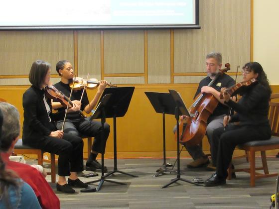 Four people playing string instruments in front of an audience.